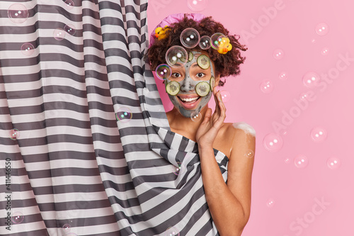 Fototapeta Naklejka Na Ścianę i Meble -  Good looking cheerful young Afro American woman applies clay mask with cucumber slices on face hides her naked body takes care of body enjoys daily hygiene routine soap bubbles flying around