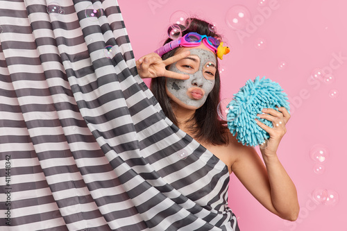 Lovely romantic Asian woman makes peace gesture over eyes keeps lips folded wears swim goggles takes shower enjoys pampering routine poses in bathroom against pink background soap bubbles around