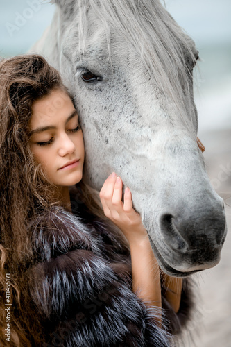 A young teenage girl in a fur coat, with long curly hair in a soft blue dress hugs a white horse on the Bank of the oken photo