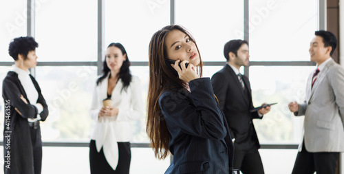 Cute Asian businesswoman using and talking on phone in front of diverse group of businesspeople.