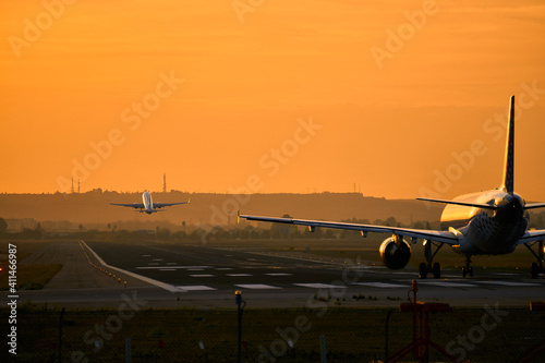 Aeropuerto con dos aviones a la vez sobre al pista de aterrizaje, uno despegando y otro esperando en un horizonte anaranjado.