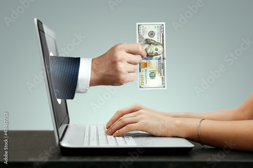A businessman's hand crawls out of a laptop screen with a hundred dollar bill. Concept of money transfers, internet banking work online.