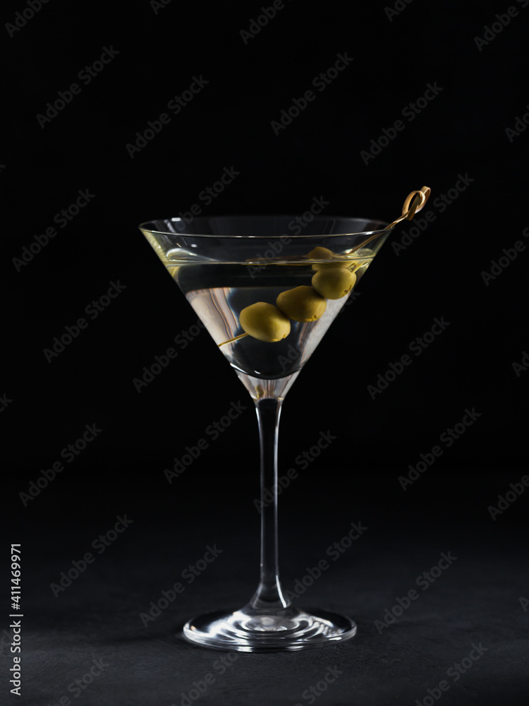 Glass of classic dry martini cocktail with olives on dark stone table against a black background.