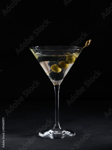 Glass of classic dry martini cocktail with olives on dark stone table against a black background.