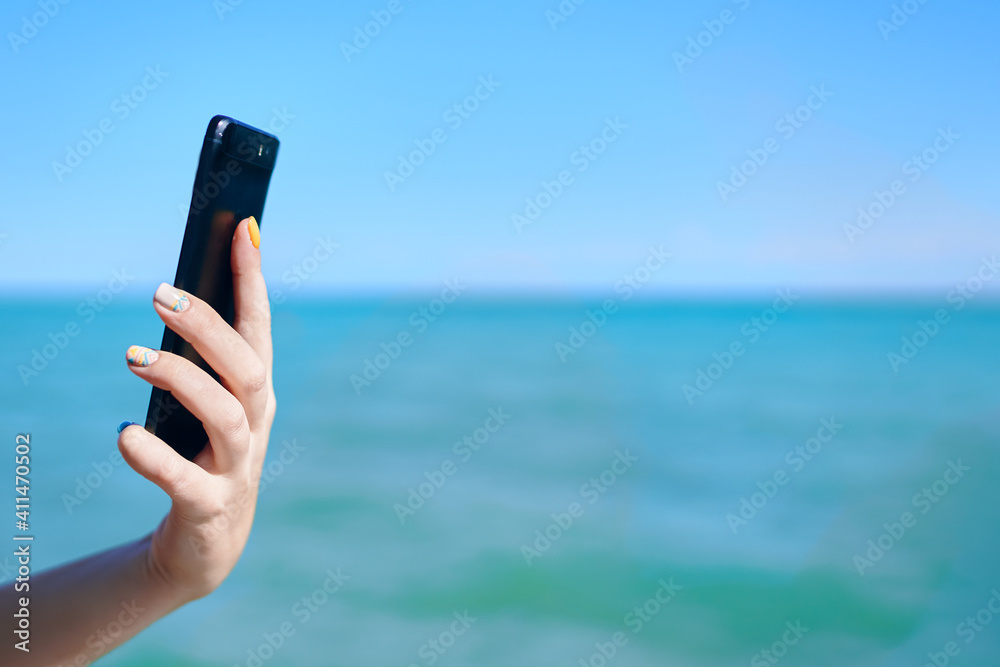 Mobile phone in woman's hand. Girl holds smartphone on sea background. Sunny summer day. Roaming and communication concept.