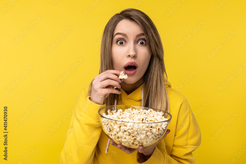 Photo of attractive lady holds plate of popcorn, eats it with shocked face. Wears casual yellow hoody, isolated yellow color background