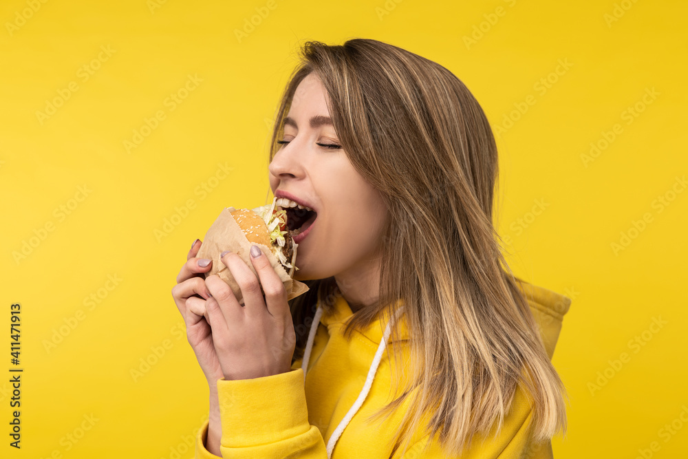Photo of attractive lady poses in a surprised way bites burger. Wears casual yellow hoody, isolated yellow color background Photo of attractive lady poses in a surprised way bites burger. Wears casual yellow hoody, isolated yellow color background