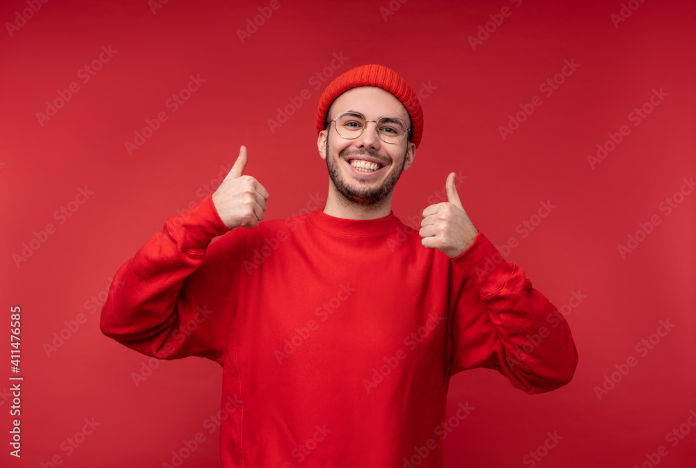 Naklejka premium Photo of attractive man with beard in glasses and red clothing. Male gives thumb up and smiles, isolated over red background