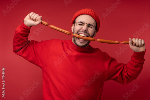 Photo of happy man with beard in glasses and red clothing. Holds and eats sausages, isolated over red background