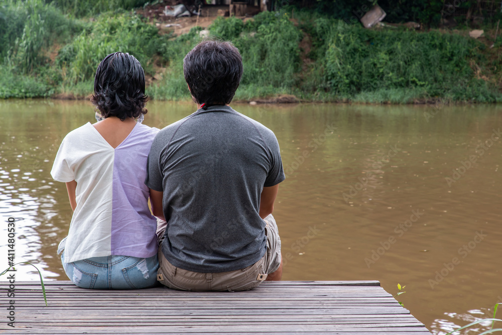 Rear view of young couple sitting in the border of a canal and watching river movement with together. Selective focus. Rear view of young couple sitting in the border of a canal and watching river movement with together. Selective focus.