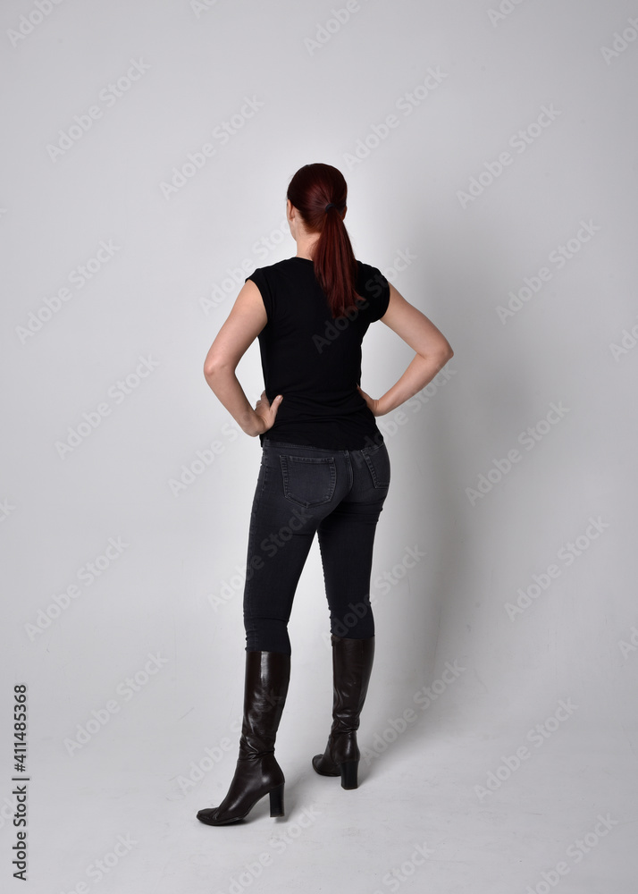 Simple full length portrait of woman with red hair in a ponytail, wearing casual black tshirt and jeans. Standing pose with back to the camera the camera, against a studio background. Simple full length portrait of woman with red hair in a ponytail, wearing casual black tshirt and jeans. Standing pose with back to the camera the camera, against a studio background.