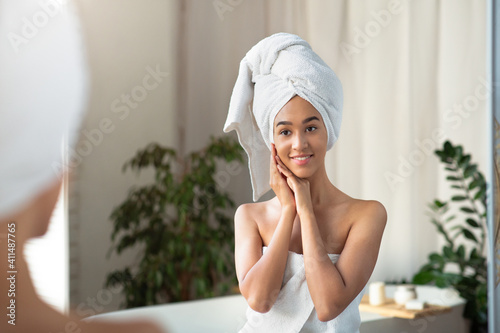 Satisfied millennial african american woman in towel presses hands to face and looks at reflection in mirror in bathroom
