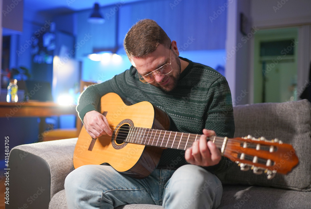 Obraz premium Young man sitting at home on sofa and playing guitar