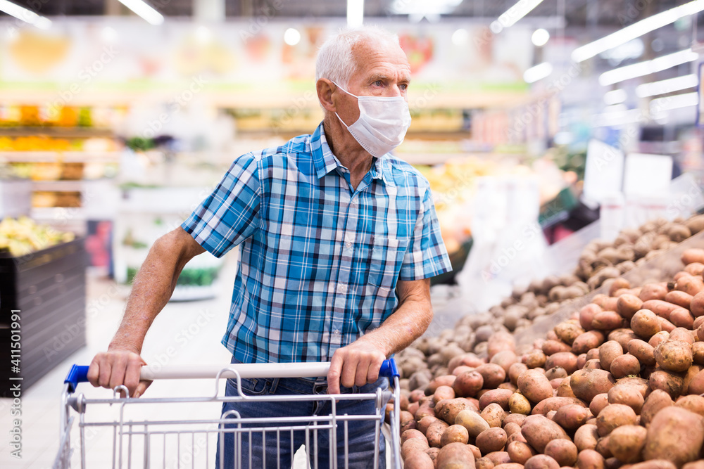 mature caucasian man in mask with covid protection choosing potatoes in vegetable section of supermarke