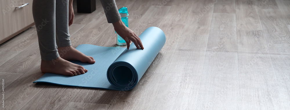 Teenage girl doing fitness exercise, practicing yoga at home. Healthy lifestyle concept. She spreads the yoga mat. Workout at home. Home interior, daytime. - obrazy, fototapety, plakaty 