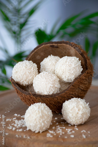 Homemade white chocolate candies in coconut flakes in nut shell on wooden board and green palm leaf. Easy recipe of tasty dessert of praline truffles bliss balls with cream. Healthy nutrition.