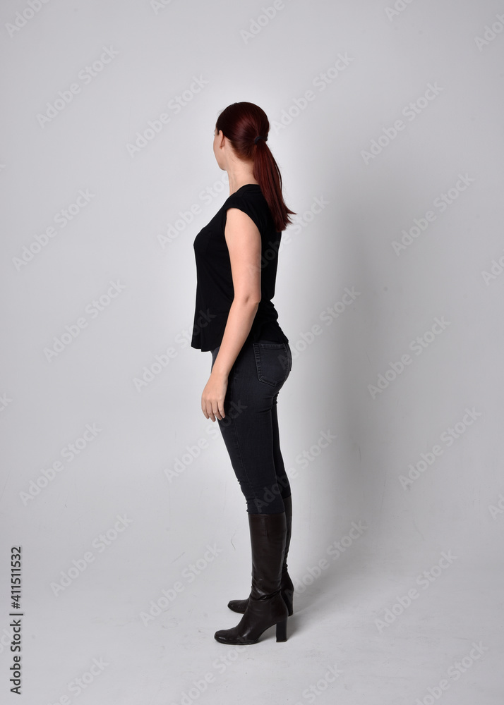 Simple full length portrait of woman with red hair in a ponytail, wearing casual black tshirt and jeans. Standing pose with in side profile, against a studio background. Simple full length portrait of woman with red hair in a ponytail, wearing casual black tshirt and jeans. Standing pose with in side profile, against a studio background.