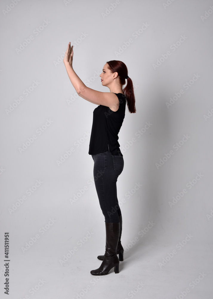 Obraz premium Simple full length portrait of woman with red hair in a ponytail, wearing casual black tshirt and jeans. Standing pose with in side profile, against a studio background.