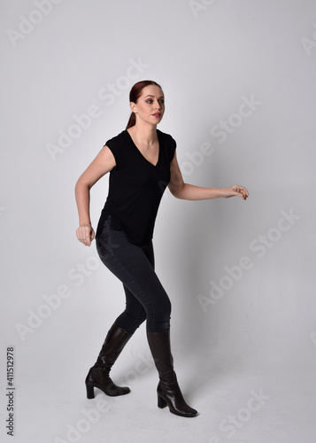 Simple full length portrait of woman with red hair in a ponytail, wearing casual black tshirt and jeans. Standing pose front on with hand reaching gestures, against a  studio background. © faestock