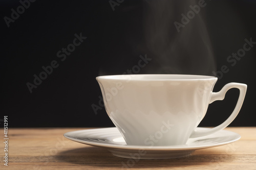 A white cup of coffee or tea with smoke on old wooden table on black background. Hot drink.