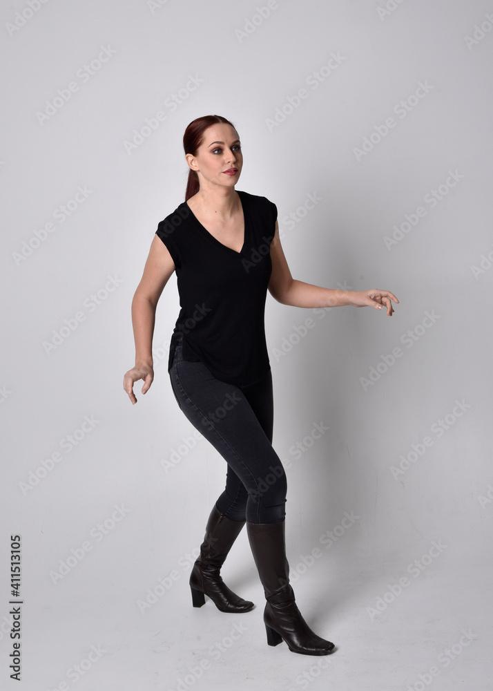 Simple full length portrait of woman with red hair in a ponytail, wearing casual black tshirt and jeans. Standing pose front on with hand reaching gestures, against a  studio background.
