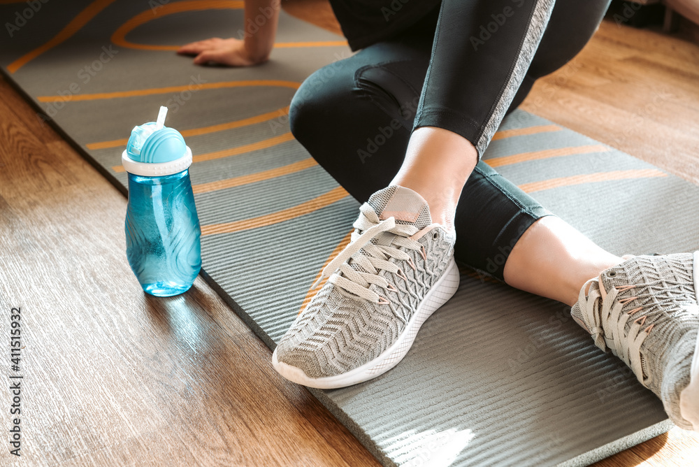 Woman sits on yoga mat rolled on the floor and drinks water from bottle resting after home fitness class, sport, active lifestyle concept - obrazy, fototapety, plakaty 