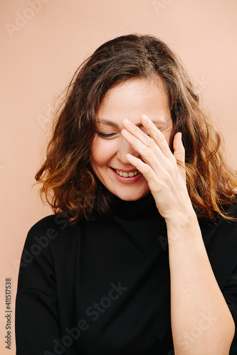 Laughing middle aged woman with her eyes closed, hand covering face. She has subtle cat eye makeup, she wears black shirt