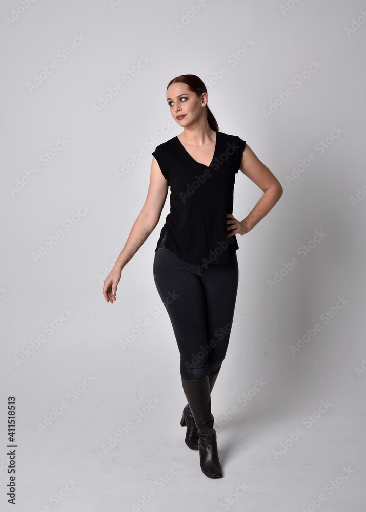 Simple full length portrait of woman with red hair in a ponytail, wearing casual black tshirt and jeans. Standing pose facing front on, against a studio background. Simple full length portrait of woman with red hair in a ponytail, wearing casual black tshirt and jeans. Standing pose facing front on, against a studio background.