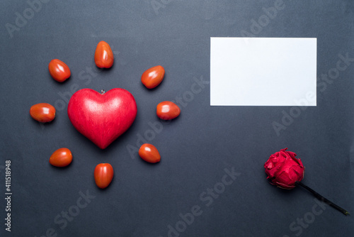 One red love heart, surrounded by small tomatoes, looking like a shining sun or a flower, with a blank gift card and a red rose next to it, black background. Concept of love and valentine's day