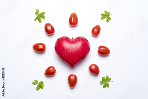 One red love heart, surrounded by small tomatoes and clover leaves, looking like a shining sun or a flower, white background. Concept of love and valentine's day