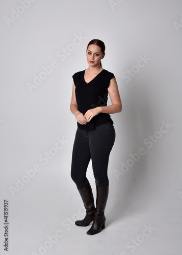 Simple full length portrait of woman with red hair in a ponytail, wearing casual black tshirt and jeans. Standing pose facing front on, against a studio background.