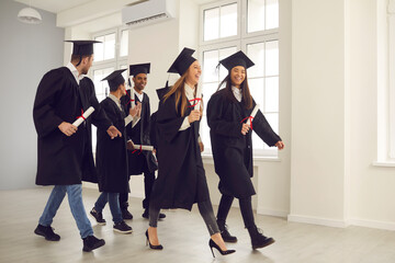 Students after the graduation ceremony. Group of happy multiracial university graduates in graduation gowns walk down the hall with diplomas in hand and communicate with each other. Success concept.