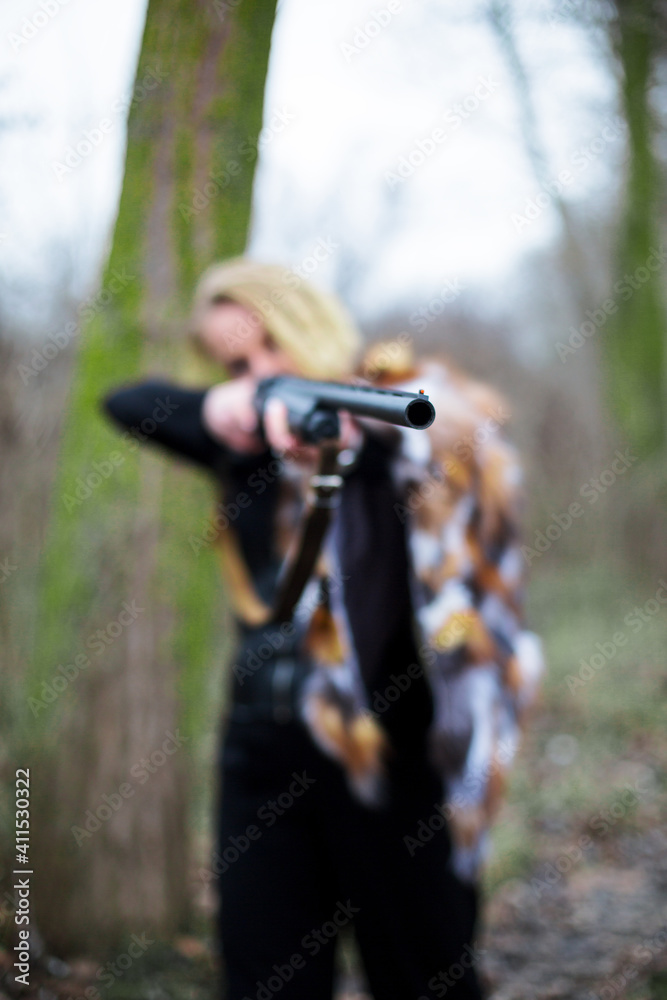 caucasian woman in black suit, corset and fur on the shoulder aim with a gun in the forest. barrel in focus