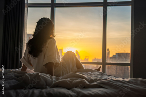 The sun shines through window in the morning. Beautiful young woman sitting on bed. Happy young girl greets new day with warm sunlight flare. Selective focus