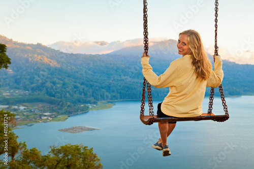 Summer vacation. Young woman sit on tree rope swing on high cliff above tropical lake. Happy girl looking at amazing jungle view. Buyan lake is popular travel destinations in Bali island, Indonesia photo