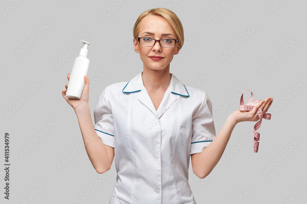 Portrait of beauty specialist standing against grey background and holding in her hand bottle of body lotion and measure tape Portrait of beauty specialist standing against grey background and holding in her hand bottle of body lotion and measure tape