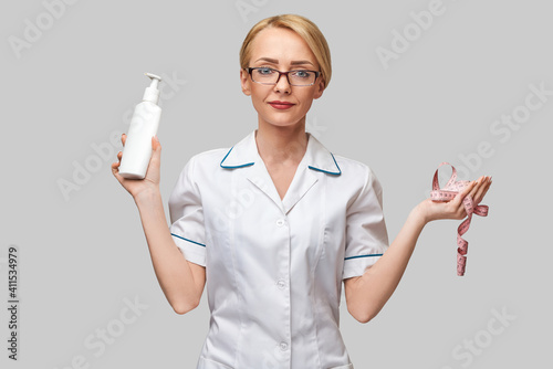 Portrait of beauty specialist standing against grey background and holding in her hand bottle of body lotion and measure tape