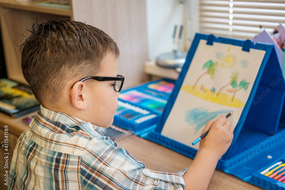 cute European boy with glasses draws on paper with pencils, sitting at the table. the process of creating a children's drawing cute European boy with glasses draws on paper with pencils, sitting at the table. the process of creating a children's drawing