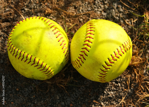 Two yellow softball close up on the ground with red lacing