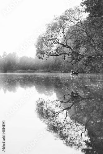 Pionowe czarno-białe ujęcie przedstawiające odbicie w tafli jeziora wędkarza na łódce i odbicie drzewa