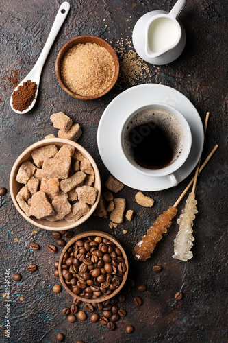 Cup of coffee with ingredients: bowls of brown cane lump and granulated sugar, crystal sugar sticks, milk and coffee beans