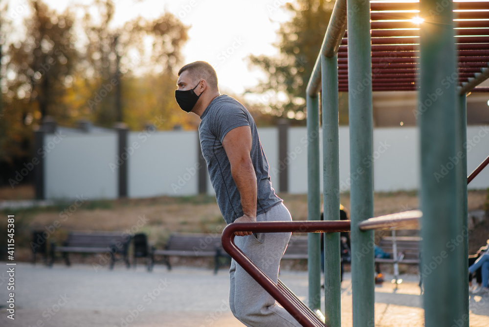 A young man does push-UPS, pull-UPS on a sports field in a mask during a pandemic at sunset. Sports, healthy lifestyle