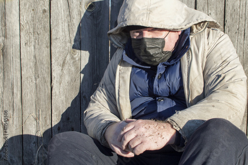 Homeless. A man in ragged clothes and a medical mask sits by a wooden fence. COVID-19.