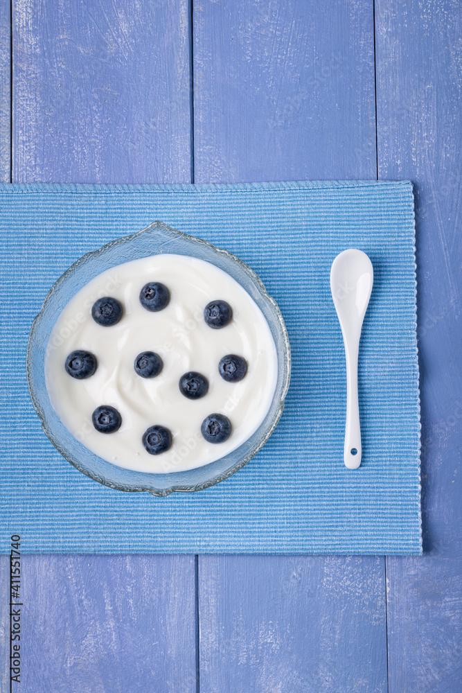 Top view, on a lavender blue background the glass bowl with low-fat white yogurt and fresh blueberries. Top view, on a lavender blue background the glass bowl with low-fat white yogurt and fresh blueberries.