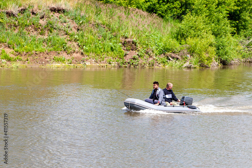Two fishermen float in an inflatable boat with an outboard motor on a summer river.