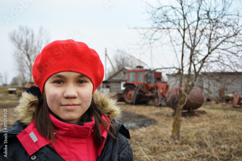 A girl in a red beret and jacket with a dirty face looks at the camera after working in the garden. A tractor stands behind her  spring  field work