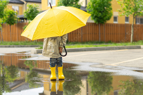 A child holds a yellow umbrella standing in a puddle outside. On sunny day happy kid walks near the house, plays, has fun