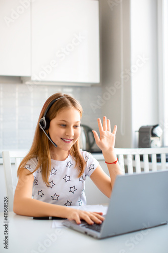 A girl in headset in front of a laptop monitor waving hand to a teacher before an online lesson. Vertical photo.