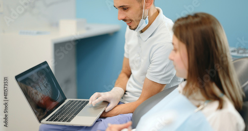 Dentist showing photo of teeth on a laptop for a young patient during an orthodontic treatment. Girl having a consultation with an orthodontist. 4k video screenshot, please use in small size