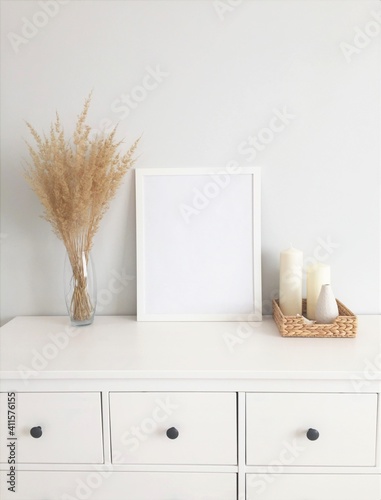 A white frame for a picture on a chest of drawers with dried herbs in a vase and white candles